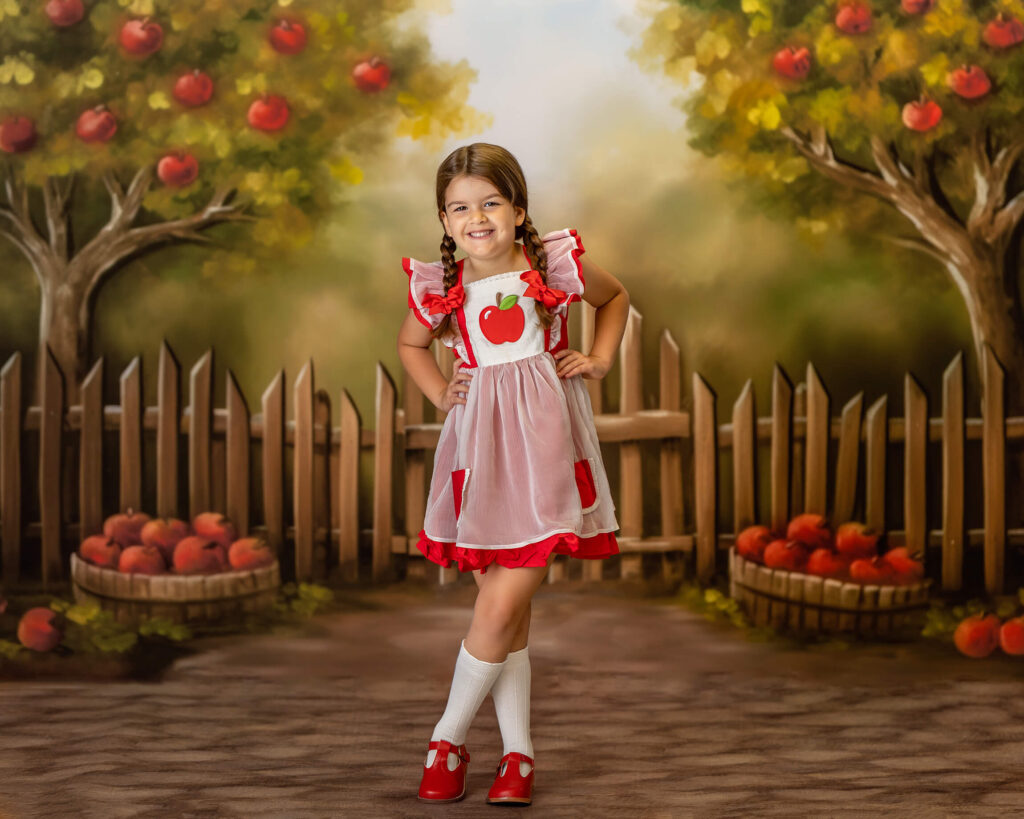 A little girl poses with apples all around her, wearing an apple dress.