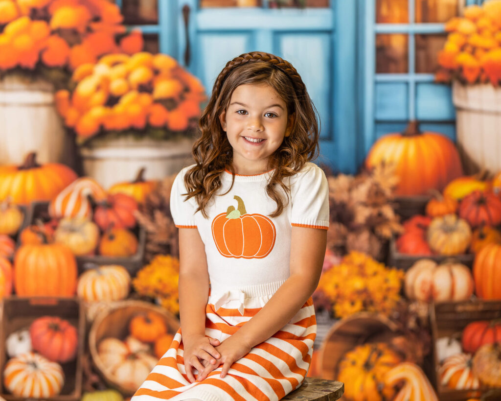 A little girl in a pumpkin sweater sits in front of a large group of pumpkins in Warwick NY