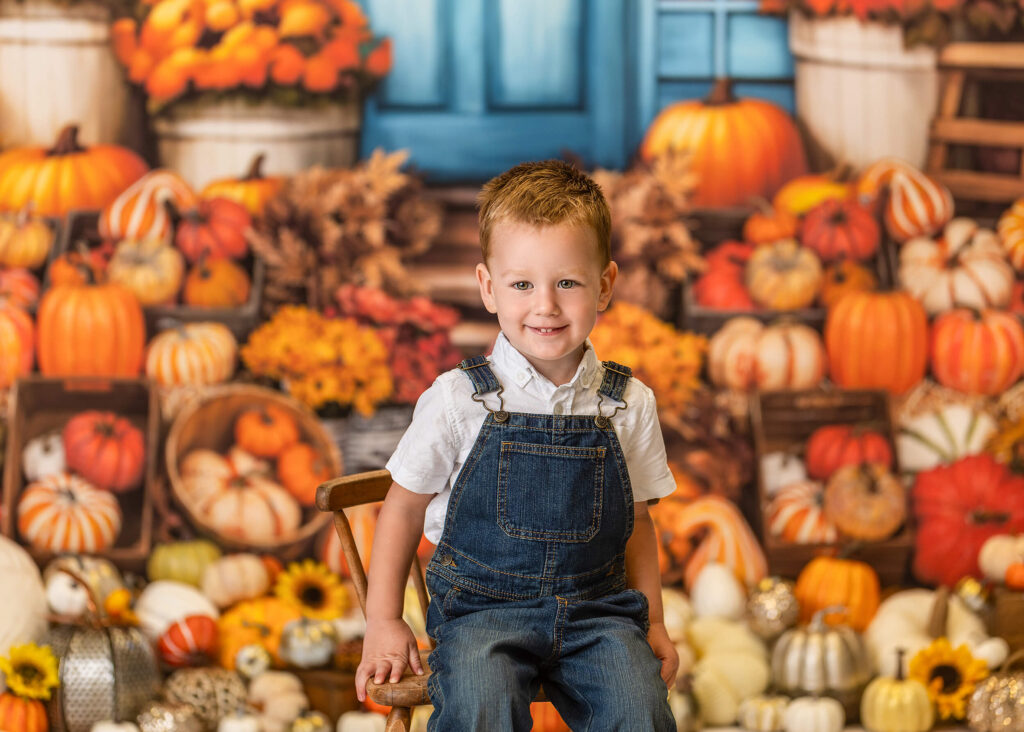 A little boy in a pair of blue overalls sits smiling in front of a large group of pumpkins in Warwick NY